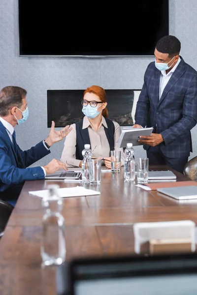 Hombre de negocios afroamericano con carpeta de papel de pie cerca del ejecutivo femenino mirando a su colega en primer plano borroso - foto de stock