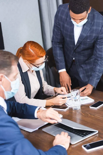 Multicultural businesspeople pointing with pens at document at workplace in boardroom with blurred colleague on foreground — Stock Photo