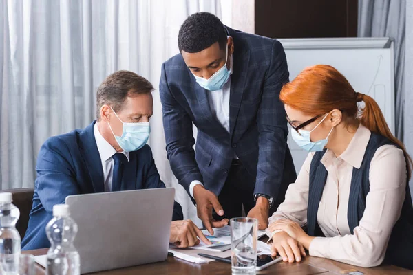 Multicultural businesspeople in medical masks pointing with fingers at document at workplace in meeting room — Stock Photo