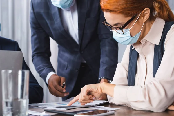 Redhead executive in medical mask pointing with finger at paper near african american businessman on blurred foreground — Stock Photo