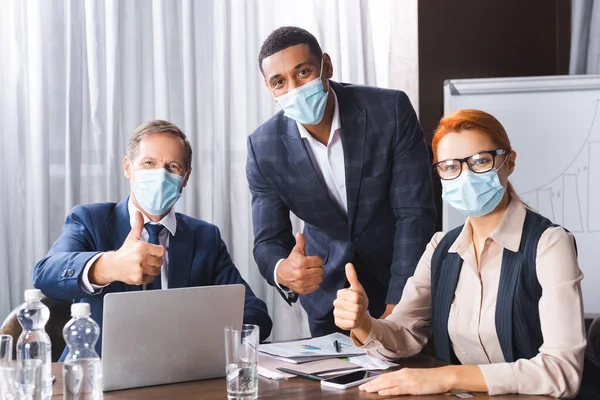 Multicultural businesspeople in medical masks looking at camera while showing thumbs up at workplace — Stock Photo