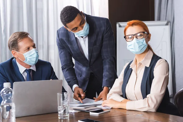 Female executive in medical mask looking at camera while sitting near multicultural colleagues in meeting room — Stock Photo