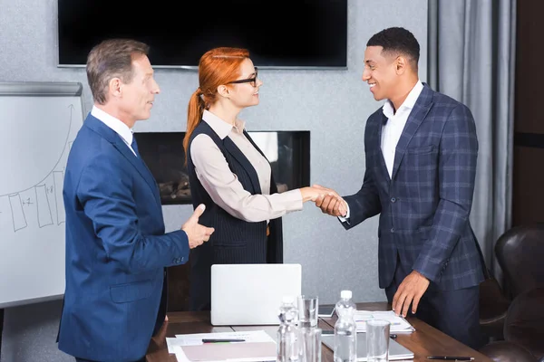Empresários multiculturais sorridentes apertando a mão uns dos outros perto de colega no local de trabalho — Fotografia de Stock