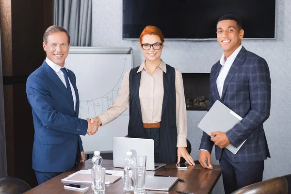 Smiling african american businessman looking at camera near colleagues shaking hands with each other in meeting room — Stock Photo