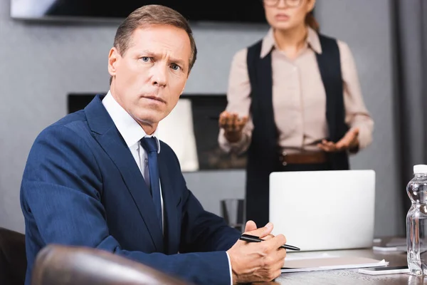 Verängstigter Geschäftsmann blickt in die Kamera, während er am Arbeitsplatz sitzt, mit verschwommener weiblicher Führungskraft im Hintergrund — Stockfoto