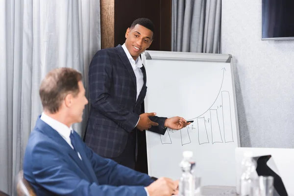 Smiling african american businessman pointing with pen at graph on flipchart and looking at blurred colleague on foreground — Stock Photo
