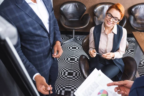 High angle view of smiling redhead executive sitting near colleagues talking near blurred flipchart on foreground — Stock Photo