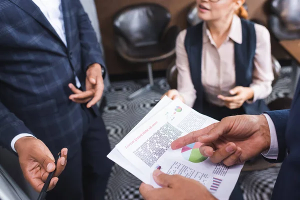 Vista recortada del hombre de negocios señalando con el dedo en el papel cerca borrosa colegas multiculturales en el fondo - foto de stock