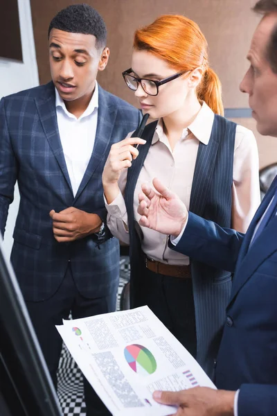 Thoughtful redhead businesswoman standing near multicultural colleagues talking and gesturing on blurred foreground — Stock Photo