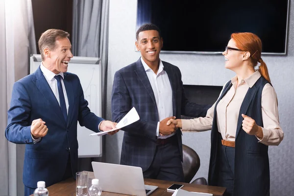 Emocionado hombre de negocios con sí gesto de pie cerca de colegas multiculturales estrechando la mano entre sí en la sala de reuniones - foto de stock