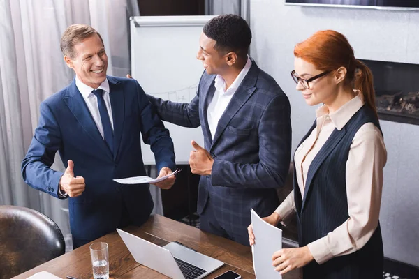 Sonrientes colegas multiculturales con los pulgares hacia arriba mirándose cerca de la pelirroja empresaria en la sala de reuniones - foto de stock