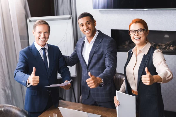 Happy multicultural businesspeople looking at camera while showing thumbs up near workplace in meeting room — Stock Photo