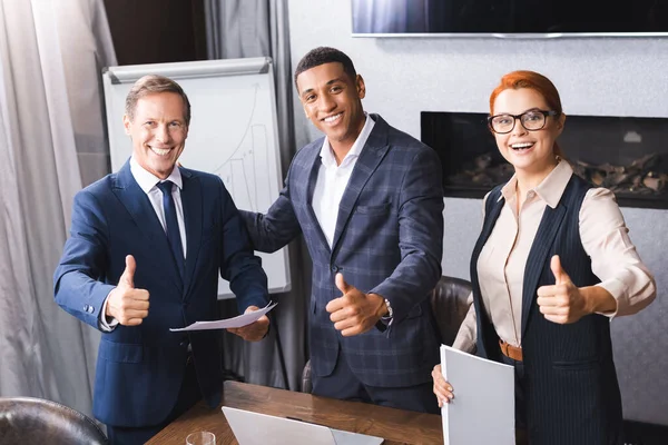 Felices empresarios multiculturales mirando la cámara mientras muestran pulgares hacia arriba cerca del lugar de trabajo en la sala de reuniones - foto de stock