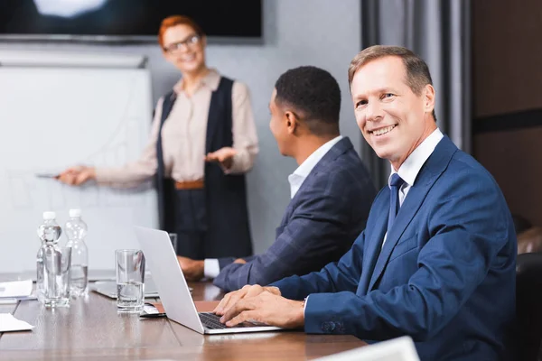 Lächelnder Geschäftsmann blickt in die Kamera, während er auf dem Laptop tippt, mit verschwommenen multikulturellen Kollegen im Hintergrund — Stockfoto