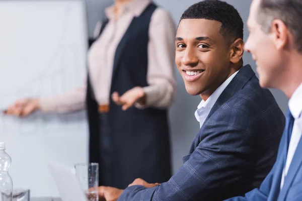 Happy african american businessman looking at camera with blurred female colleague on background — Stock Photo