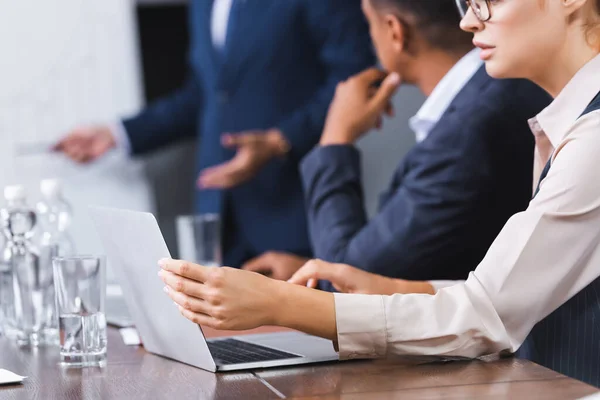 Cropped view of businesswoman using laptop while sitting at workplace with blurred multicultural colleagues on background — Stock Photo