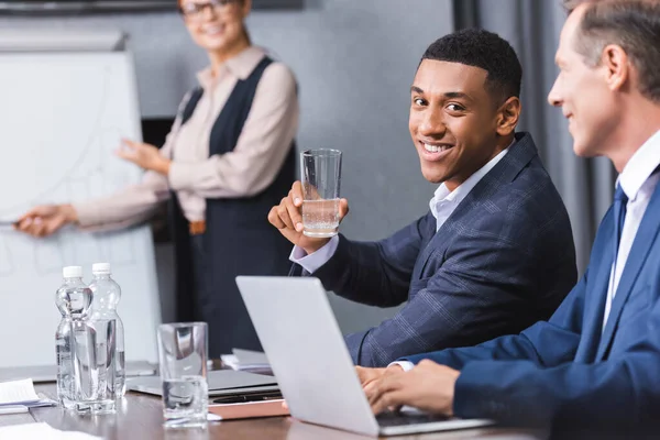 Homme d'affaires afro-américain souriant avec un verre d'eau regardant la caméra tout en étant assis sur le lieu de travail sur un fond flou — Photo de stock