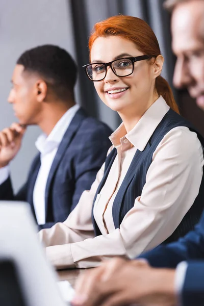 Cheerful redhead businesswoman in eyeglasses looking at camera with blurred african american colleague on background — Stock Photo