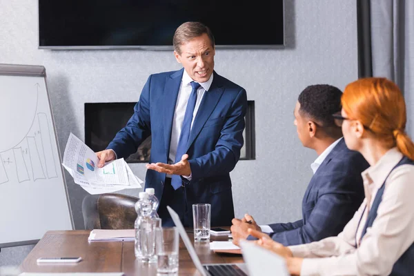 Ejecutivo enojado con papeles gritando mientras mira a los empresarios multiculturales en primer plano borroso en la sala de juntas - foto de stock