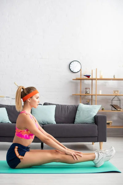 Seitenansicht einer jungen Sportlerin, die sich auf einer Fitnessmatte in der Nähe der heimischen Couch dehnt — Stockfoto