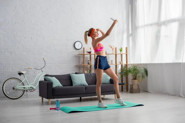 Joven deportista rubia con labios de puchero tomando selfie mientras está de pie en la colchoneta de fitness en casa — Stock Photo