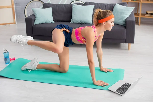 Smiling blonde sportive faisant de l'exercice sur tapis de fitness près d'un ordinateur portable à la maison — Photo de stock