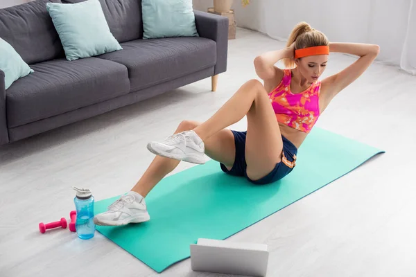 Mujer rubia joven haciendo abdominales en la alfombra de fitness cerca de la computadora portátil, mancuernas y botella de deportes en casa - foto de stock