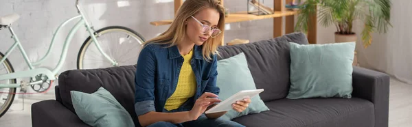 Giovane donna bionda in occhiali con tablet digitale mentre si siede sul divano a casa su sfondo sfocato, banner — Foto stock