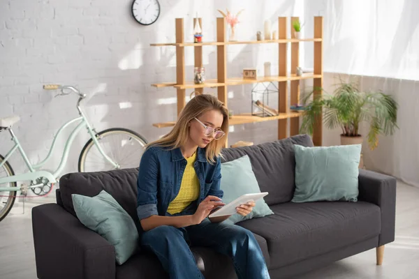 Young blonde woman in eyeglasses using digital tablet while sitting on couch at home — Stock Photo