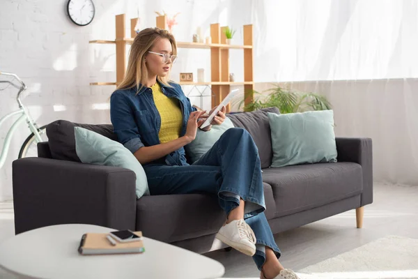 Mujer rubia joven con gafas de vista usando tableta digital mientras está sentada en el sofá en casa sobre un fondo borroso — Stock Photo