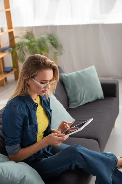 Mujer rubia joven con gafas de vista usando tableta digital mientras está sentada en el sofá sobre fondo borroso - foto de stock