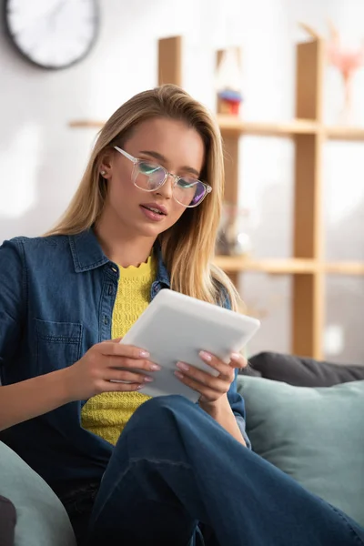 Mujer rubia con gafas de vista mirando la tableta digital mientras está sentada en el sofá sobre un fondo borroso - foto de stock