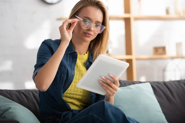 Молода блондинка торкається окулярів, дивлячись на цифровий планшет вдома на розмитому фоні — стокове фото
