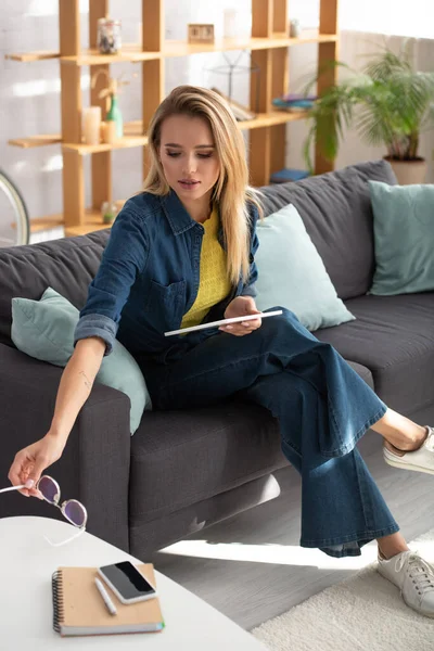 Mujer rubia joven con tableta digital poniendo gafas en la mesa de café mientras está sentado en el sofá sobre fondo borroso - foto de stock