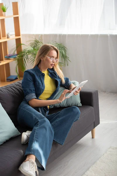 Full length of young blonde woman in eyeglasses using digital tablet while sitting on couch on blurred background — Stock Photo