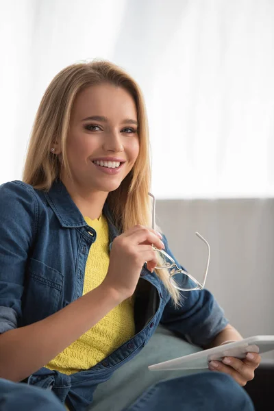 Happy blonde woman looking at camera while holding digital tablet and eyeglasses at home — Stock Photo