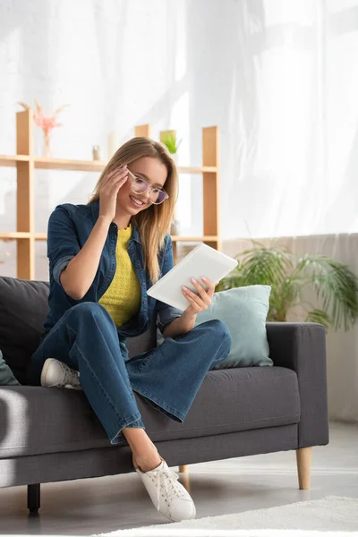 Comprimento total de sorrir jovem mulher em óculos olhando para tablet digital no sofá no fundo borrado — Fotografia de Stock