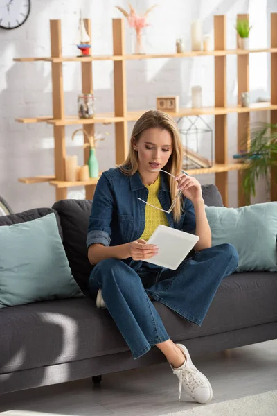 Longitud completa de mujer rubia joven con gafas mirando a la tableta digital mientras está sentado en el sofá sobre un fondo borroso - foto de stock