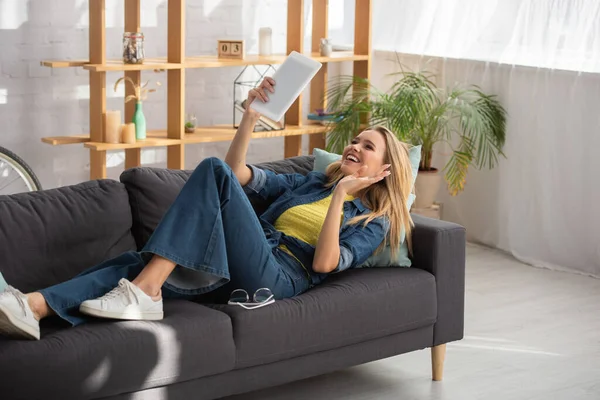 Усміхнена блондинка з махаючою рукою дивиться на цифровий планшет, лежачи на дивані вдома — стокове фото