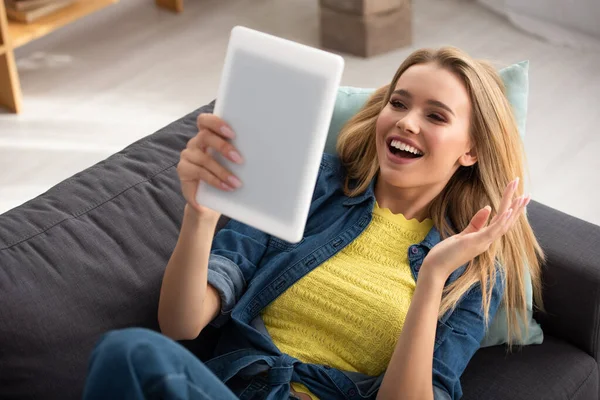 Allegro donna bionda con mano ondulata guardando tablet digitale mentre si trova sul divano su sfondo sfocato — Foto stock