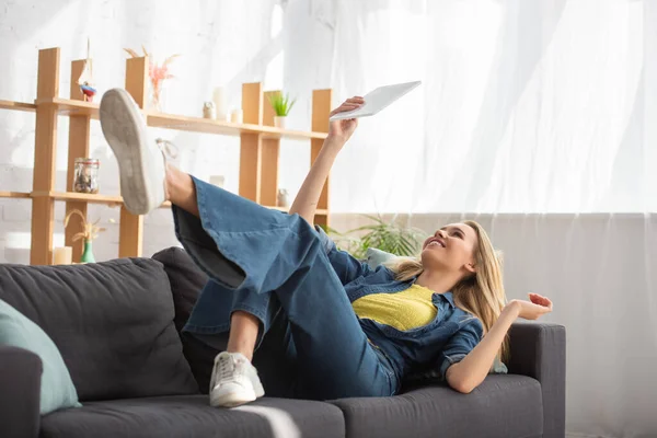 Giovane donna bionda guardando tablet digitale mentre sdraiato sul divano a casa su sfondo sfocato — Foto stock