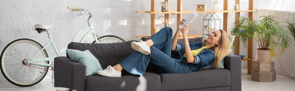 Smiling young woman looking at digital tablet during video call on couch at home, banner — Stock Photo