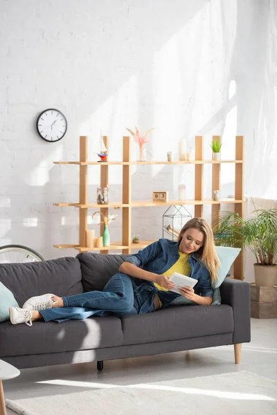 Full length of smiling blonde woman looking at digital tablet while lying on couch at home — Stock Photo