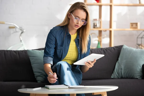 Junge blonde Frau mit digitalem Tablet schreibt in Notizbuch, während sie auf der Couch auf verschwommenem Hintergrund sitzt — Stockfoto