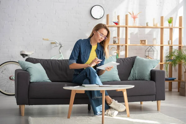 Junge Frau nutzt digitales Tablet in voller Länge, während sie zu Hause auf der Couch in der Nähe des Couchtisches sitzt — Stockfoto
