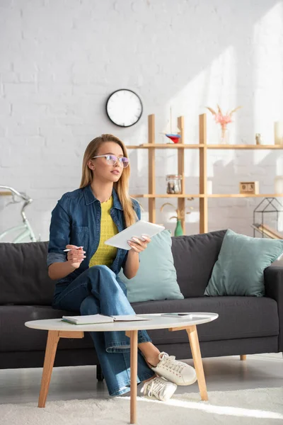 Pleine longueur de jeune femme blonde avec stylo et tablette numérique regardant loin tout en étant assis sur le canapé sur fond flou — Photo de stock