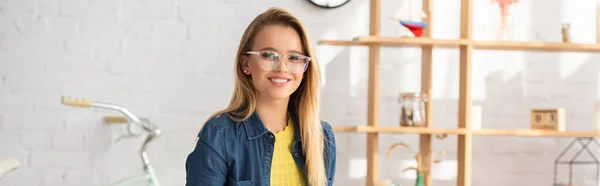 Mujer rubia alegre en anteojos mirando a la cámara en casa sobre fondo borroso, pancarta - foto de stock