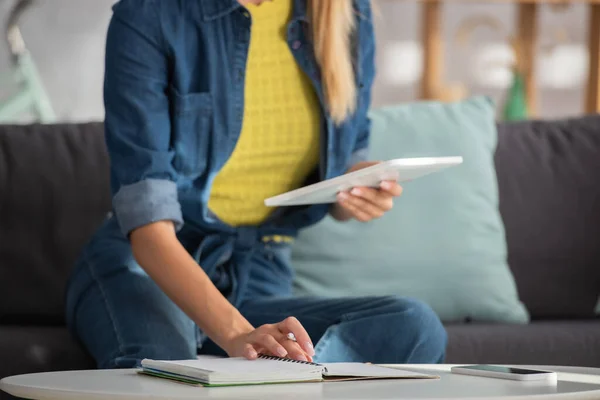 Vue recadrée de la jeune femme avec tablette numérique assise près du portable à la maison sur fond flou — Photo de stock