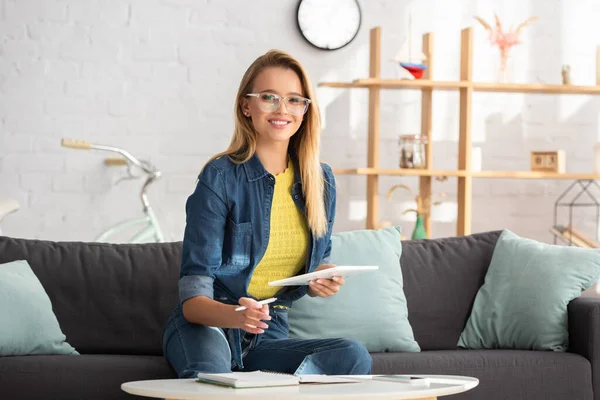 Fröhliche junge Frau mit Stift und digitalem Tablet, die auf der Couch vor verschwommenem Hintergrund in die Kamera blickt — Stockfoto