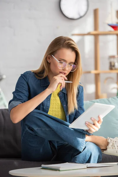 Femme blonde réfléchie avec stylo regardant tablette numérique tout en étant assis sur le canapé sur fond flou — Photo de stock